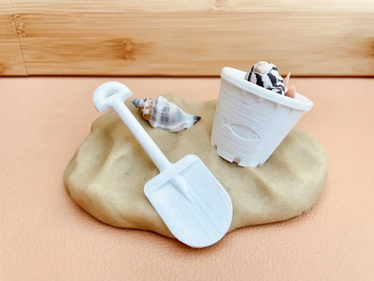 Lump of light-colored clay or dough with a miniature white shovel and a small pot containing tiny rocks or pebbles.