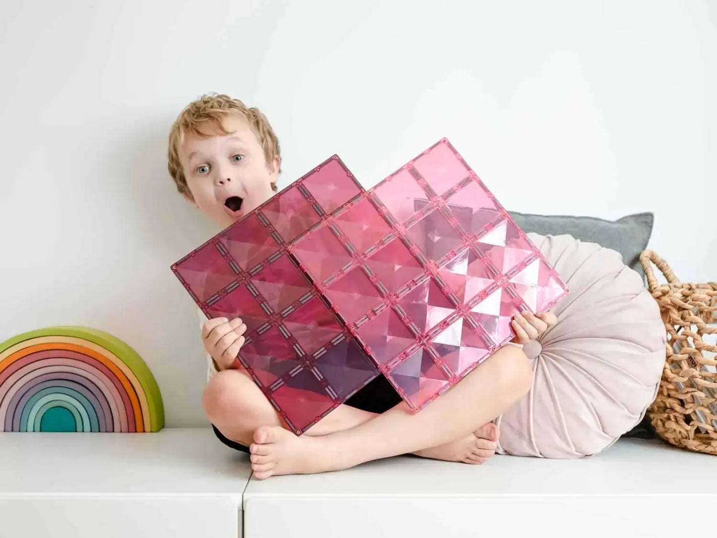 Young child holding a pink geometric cushion with a surprised expression.