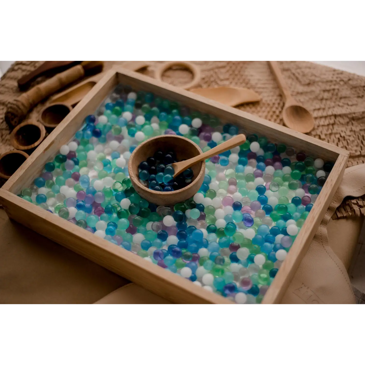 Wooden tray filled with colorful beads or marbles, containing a small bowl and wooden scoop.