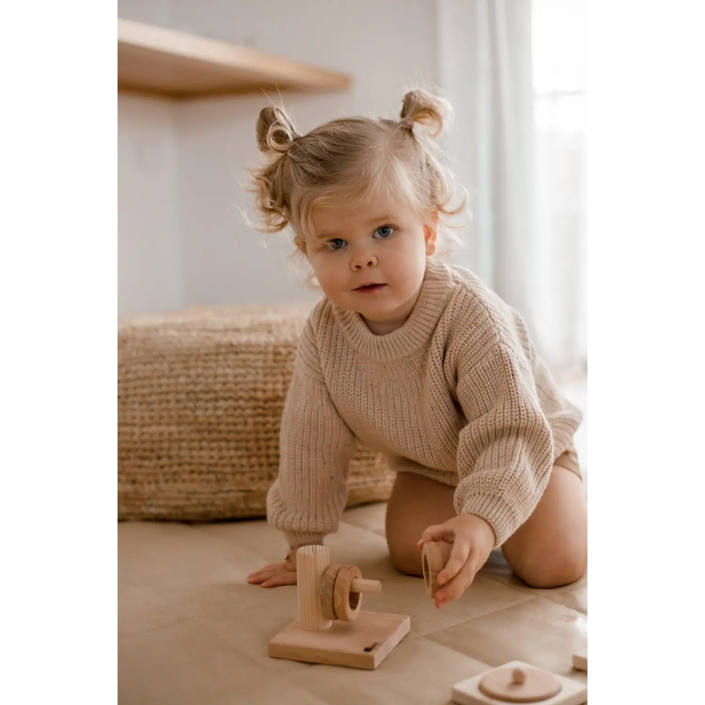 Young child with pigtails wearing a beige sweater, sitting on the floor with a wooden toy.