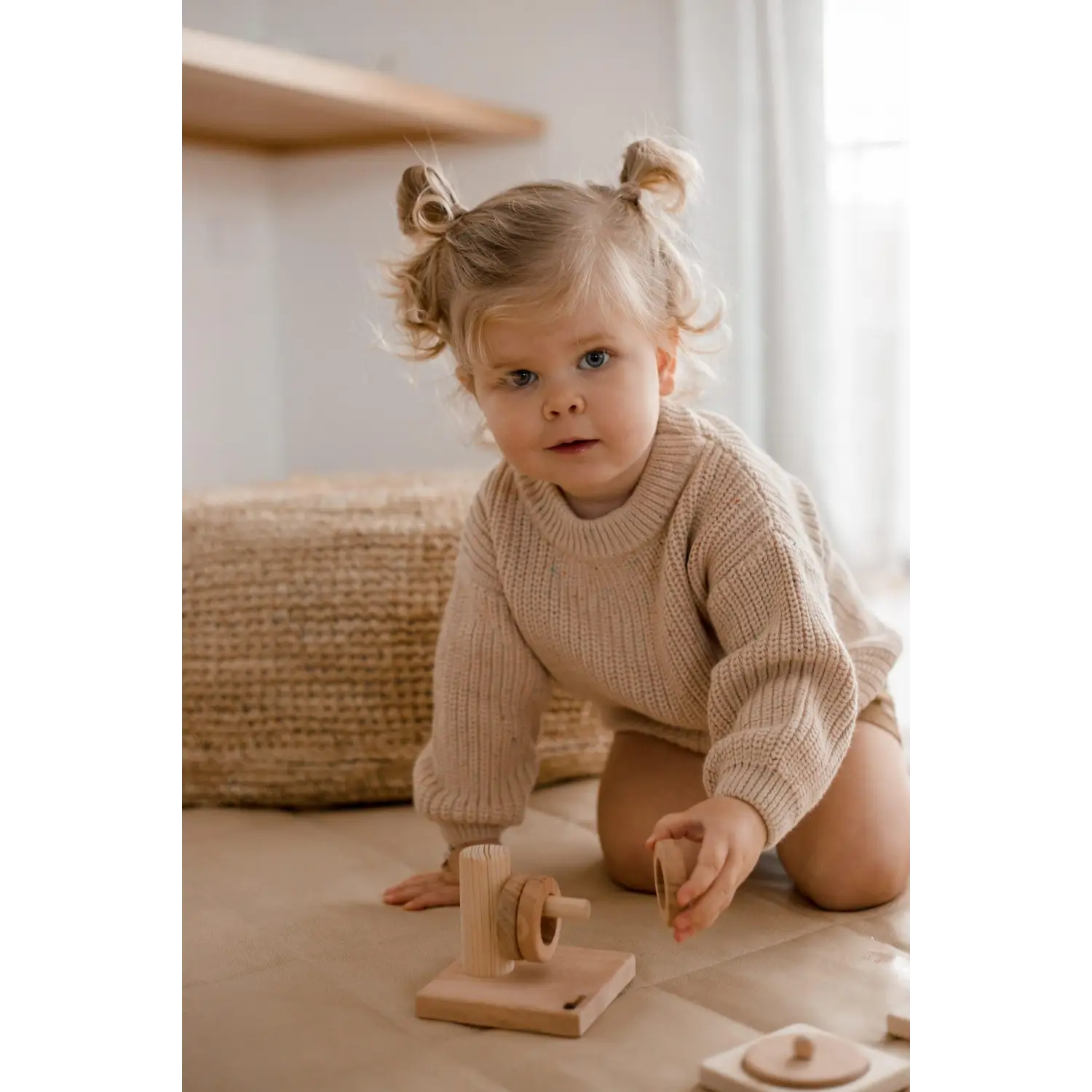 Young child with pigtails wearing a beige sweater, sitting on the floor with a wooden toy.