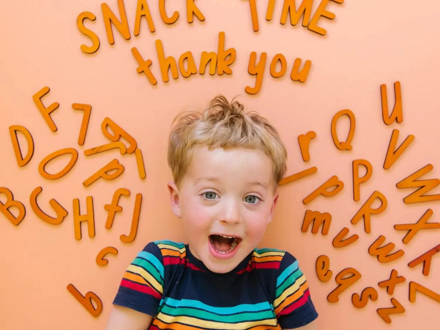 Excited young child wearing a colorful striped shirt surrounded by orange letter shapes spelling out words like ’Snack Time’ and ’thank you’.