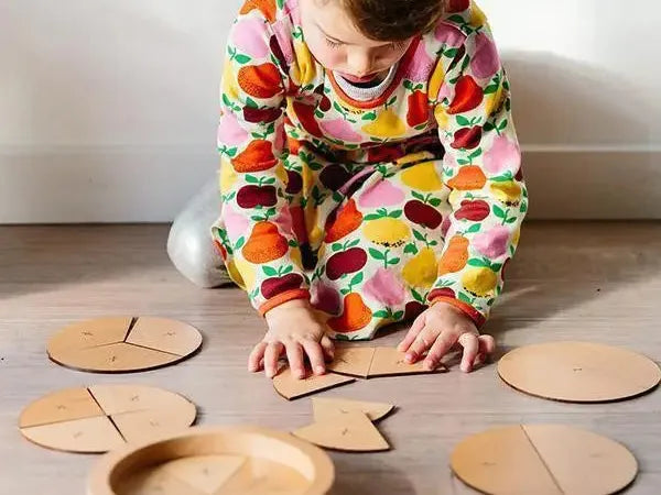 Child wearing colorful fruit-patterned pajamas playing with wooden circular puzzle pieces on the floor.