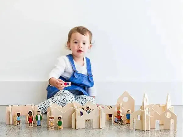 Toddler wearing blue overalls sitting behind wooden toy buildings and figurines.