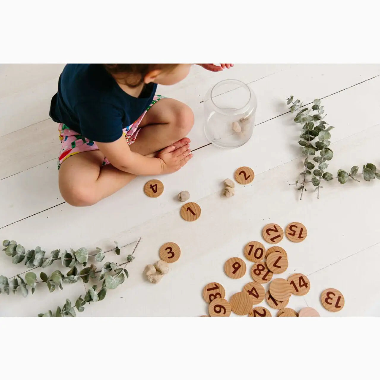Wooden number tokens scattered on a white floor alongside eucalyptus branches.