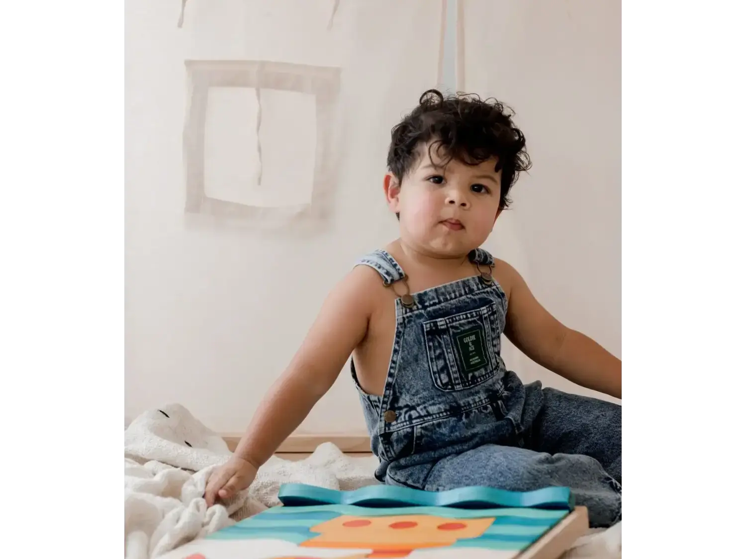 Child wearing denim overalls sitting on the floor with a colorful board game or puzzle.