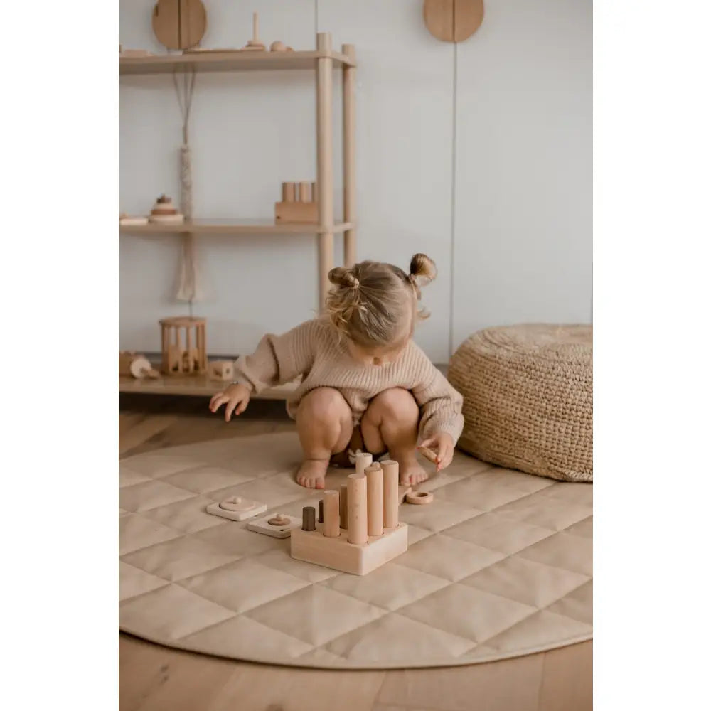 Toddler with hair buns playing with wooden blocks on a circular mat.