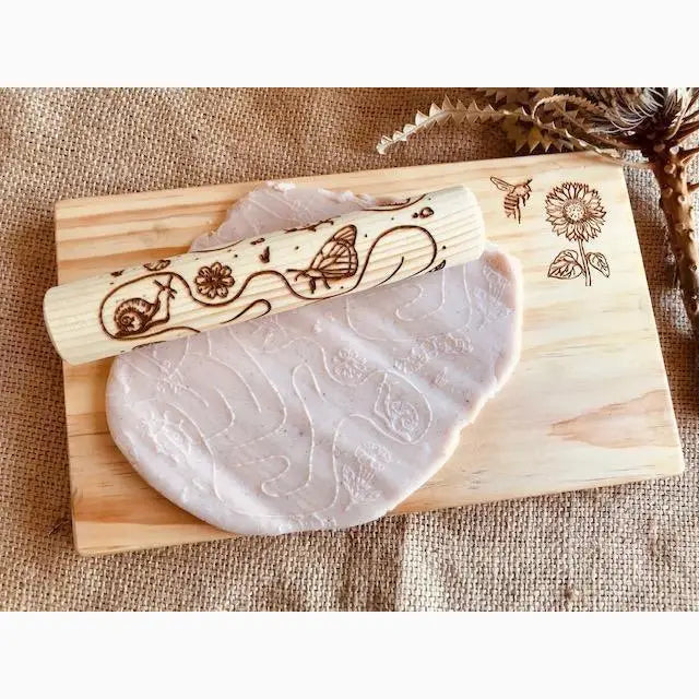 Wooden rolling pin with engraved nature-inspired designs next to a flattened piece of clay or dough.