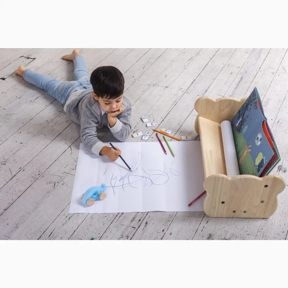 Child lying on a wooden floor drawing on a large sheet of paper.