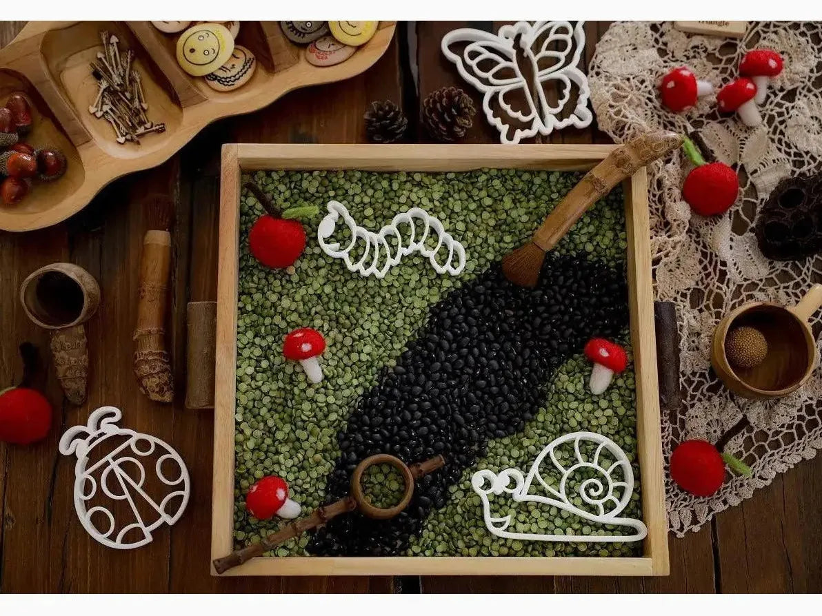 Wooden tray filled with green and black beans arranged in a decorative pattern, adorned with small red mushrooms and white ornaments.