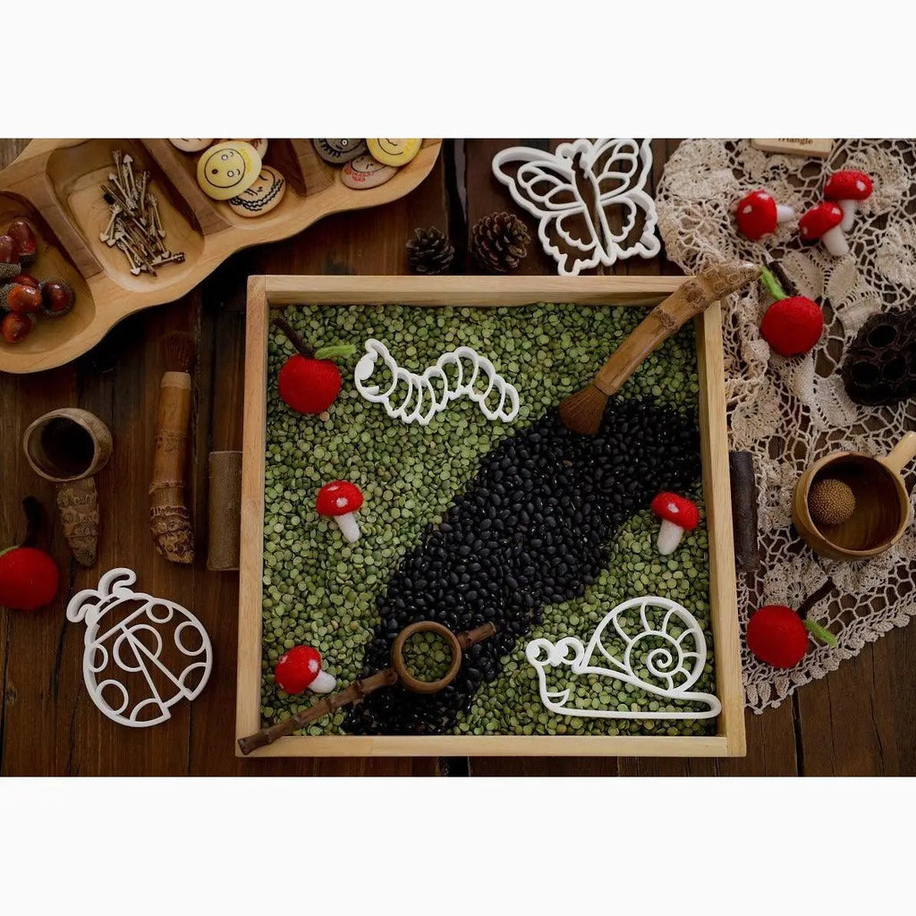 Wooden tray filled with green and black beans arranged in a decorative pattern, adorned with small red mushrooms and white ornaments.