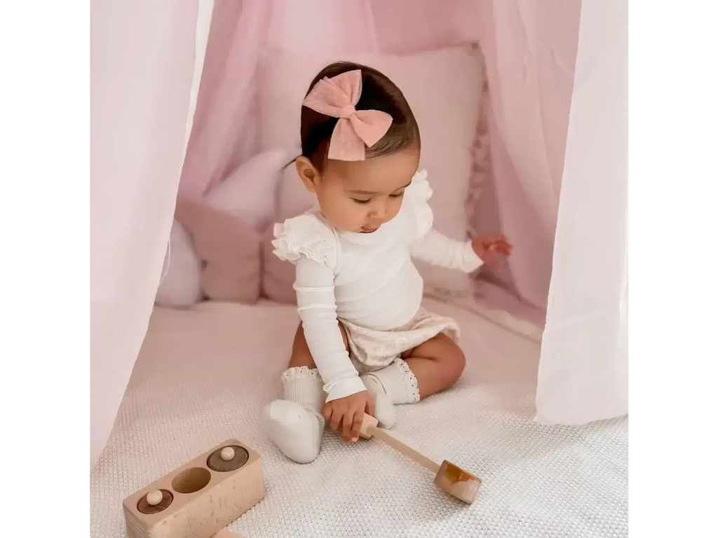 Baby wearing a white outfit and pink bow, sitting with wooden toys.