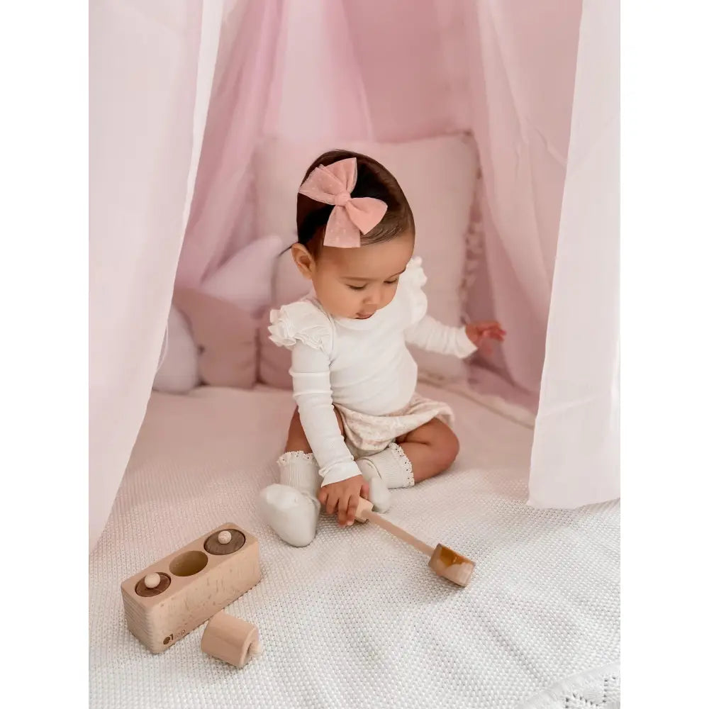 Baby wearing a white outfit and pink bow, sitting with wooden toys.