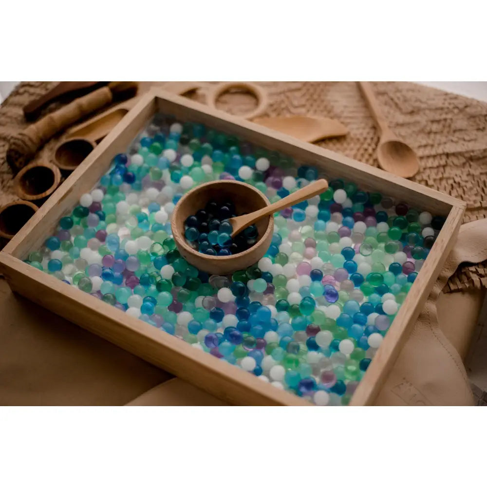 Wooden tray filled with colorful beads or marbles, containing a small bowl and wooden scoop.