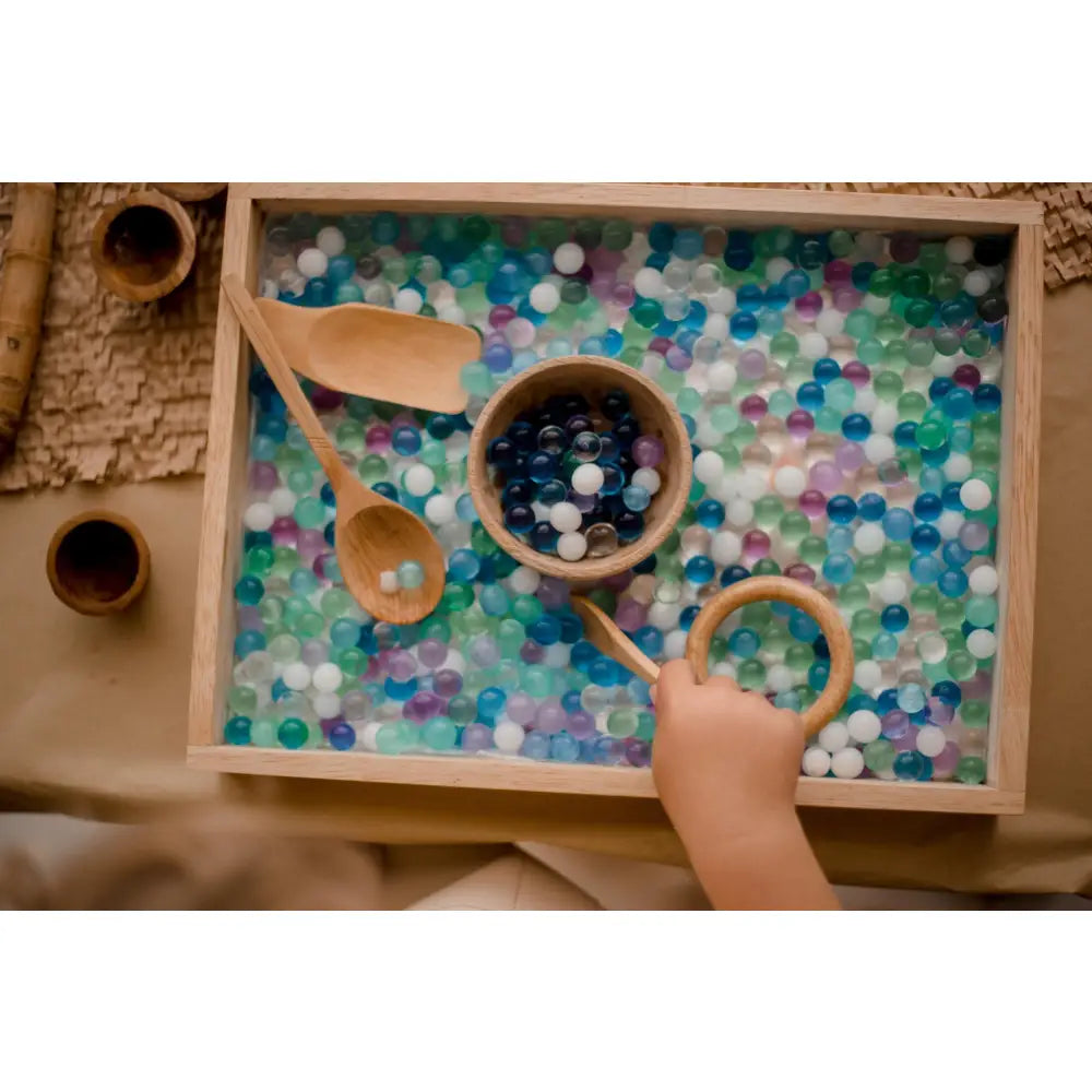 Sensory tray filled with colorful water beads and wooden scoops.