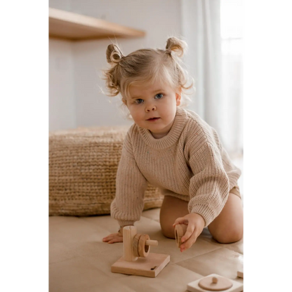 Young child with pigtails wearing a beige sweater, sitting on the floor with a wooden toy.