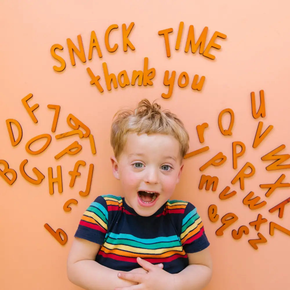 Excited young child wearing a colorful striped shirt surrounded by orange letter shapes spelling out words like ’Snack Time’ and ’thank you’.