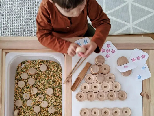 Child engaged in a sensory or educational activity with wooden discs and a tray of seeds or grains.