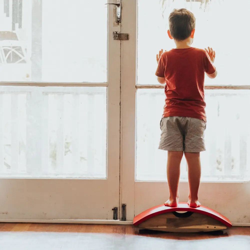 Child standing on a balance board in front of a glass door.