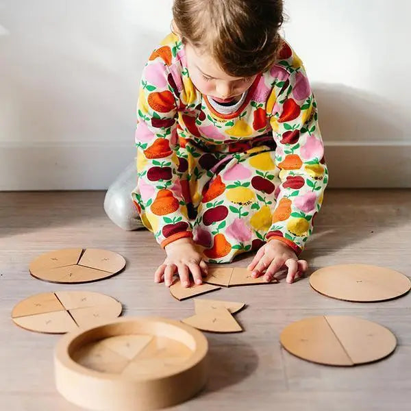 Child wearing colorful fruit-patterned pajamas playing with wooden circular puzzle pieces on the floor.