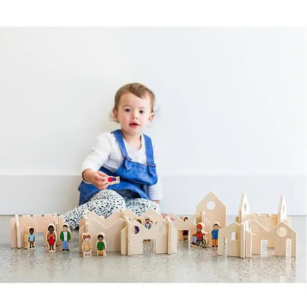 Toddler wearing blue overalls sitting behind wooden toy buildings and figurines.