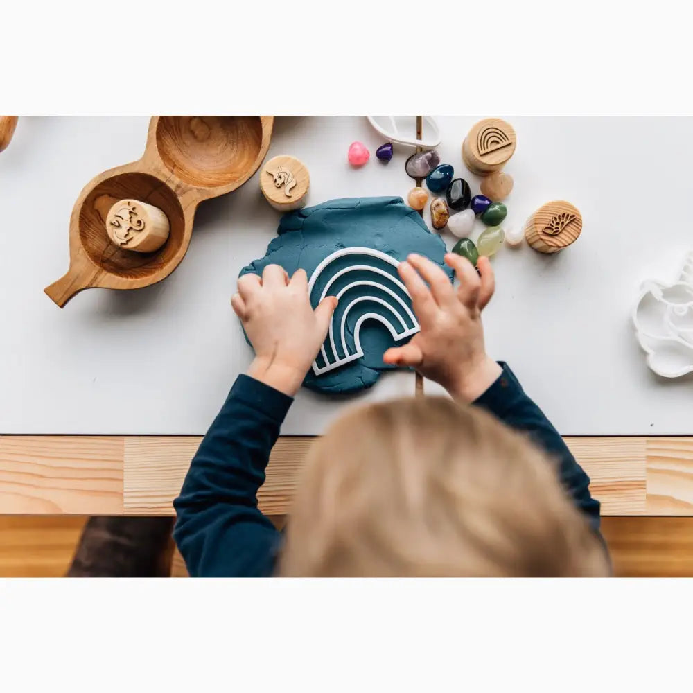 Rainbow-shaped wooden toy being held by small hands on a table with various craft materials.