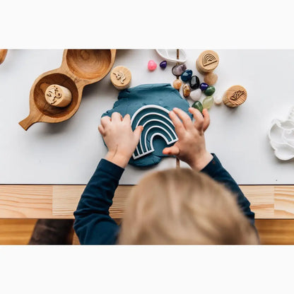 Rainbow-shaped wooden toy being held by small hands on a table with various craft materials.