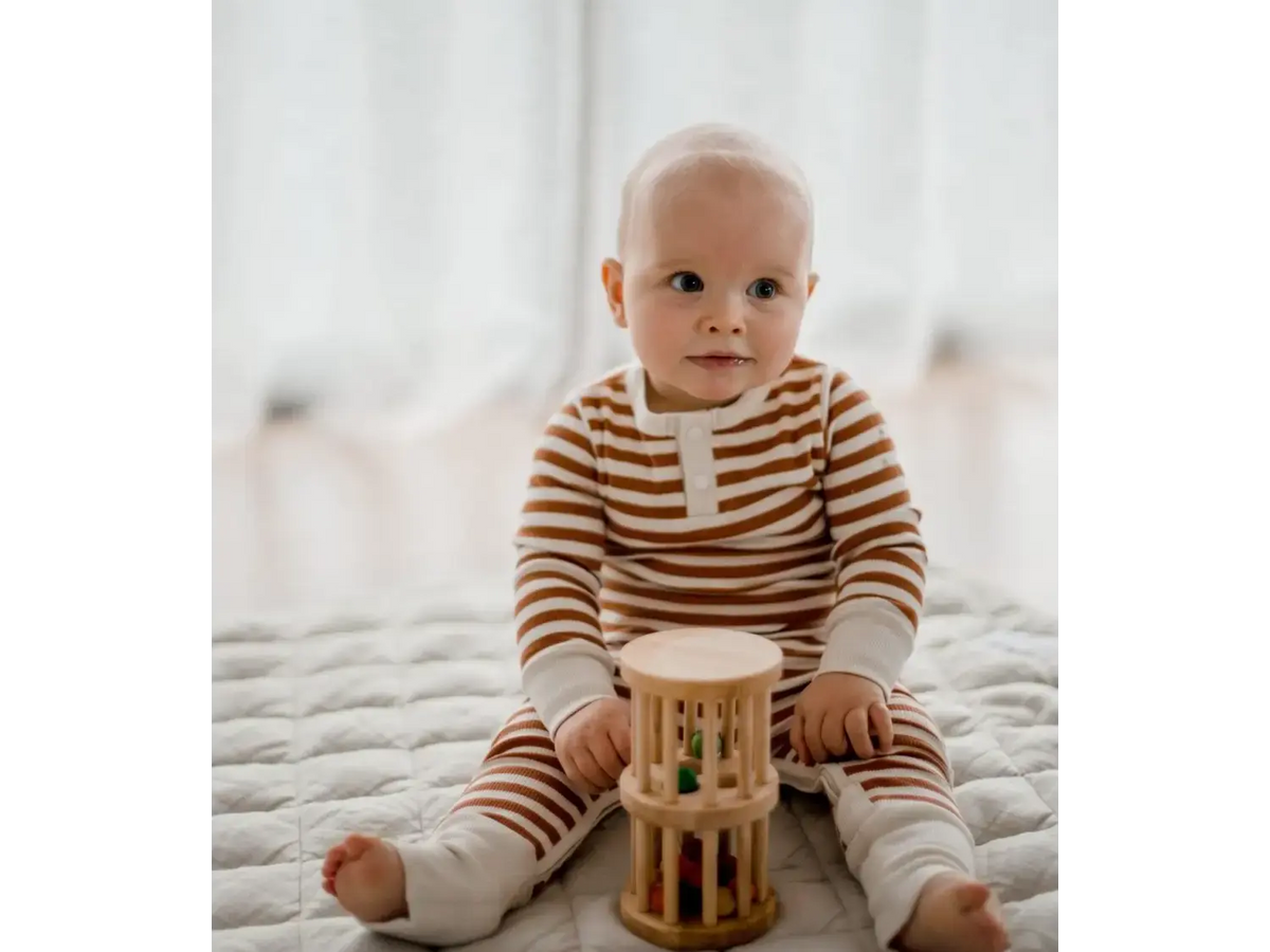 Baby wearing striped clothing sitting on a bed with a wooden toy.