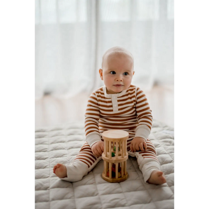 Baby wearing striped clothing sitting on a bed with a wooden toy.