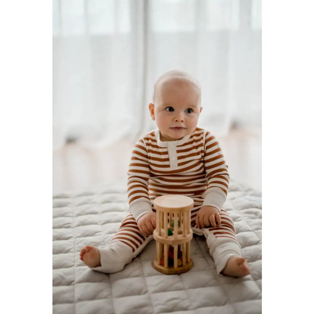 Baby wearing striped clothing sitting on a bed with a wooden toy.