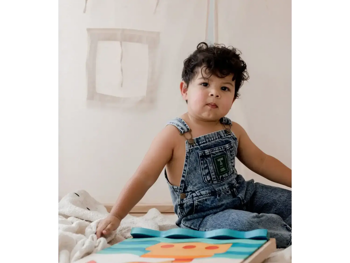 Child wearing denim overalls sitting on the floor with a colorful board game or puzzle.
