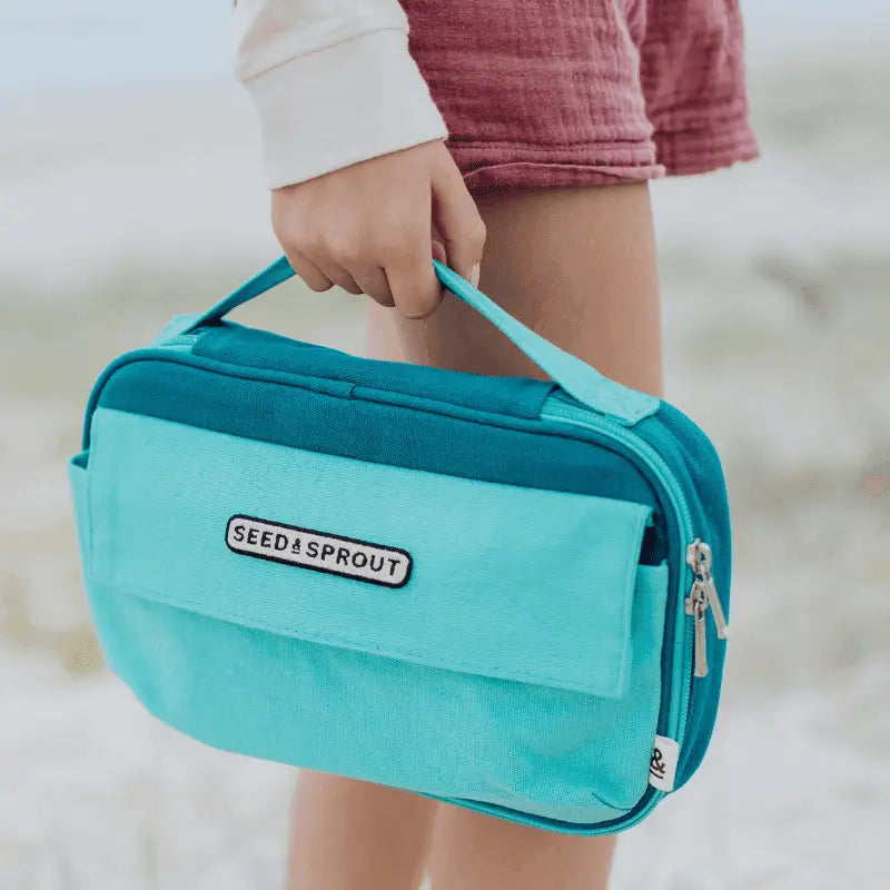 Teal-colored insulated lunch bag with ’SEED & SPROUT’ branding being carried by a hand.