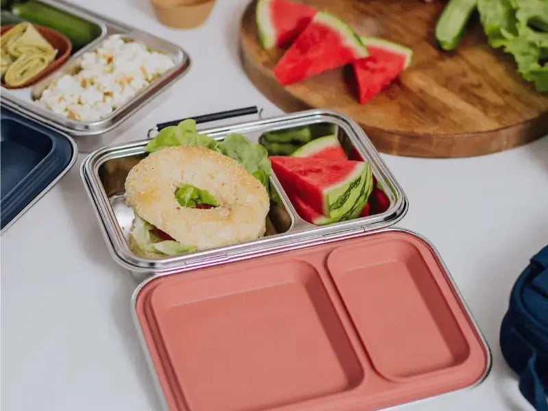 Compartmentalized lunch containers filled with various foods like watermelon, sandwich, and rice.