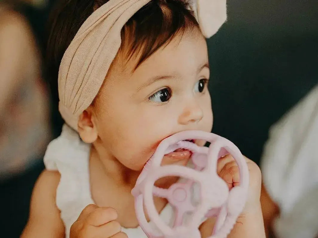 Baby wearing a cream headband and chewing on a pink teething toy.