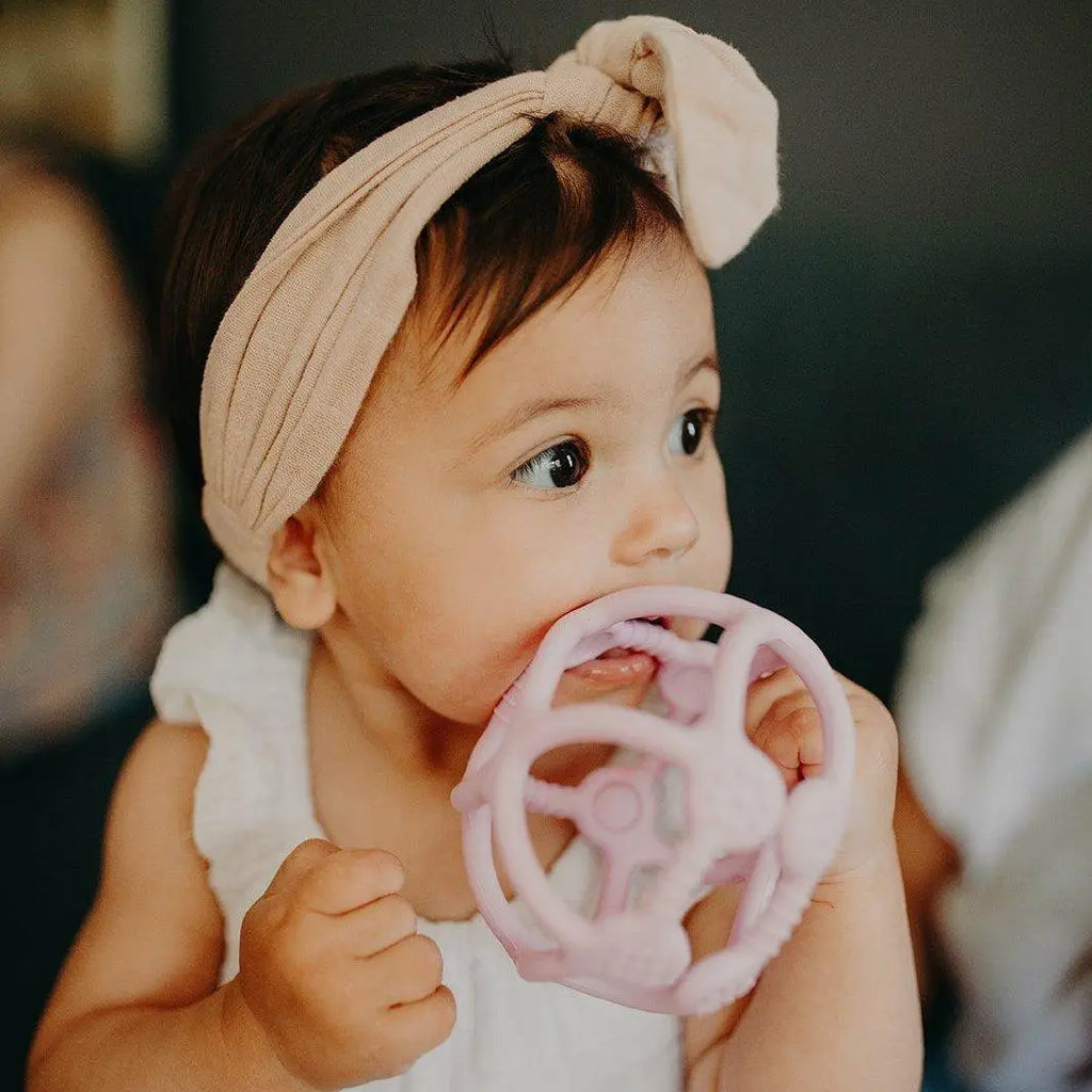 Baby wearing a cream headband and chewing on a pink teething toy.