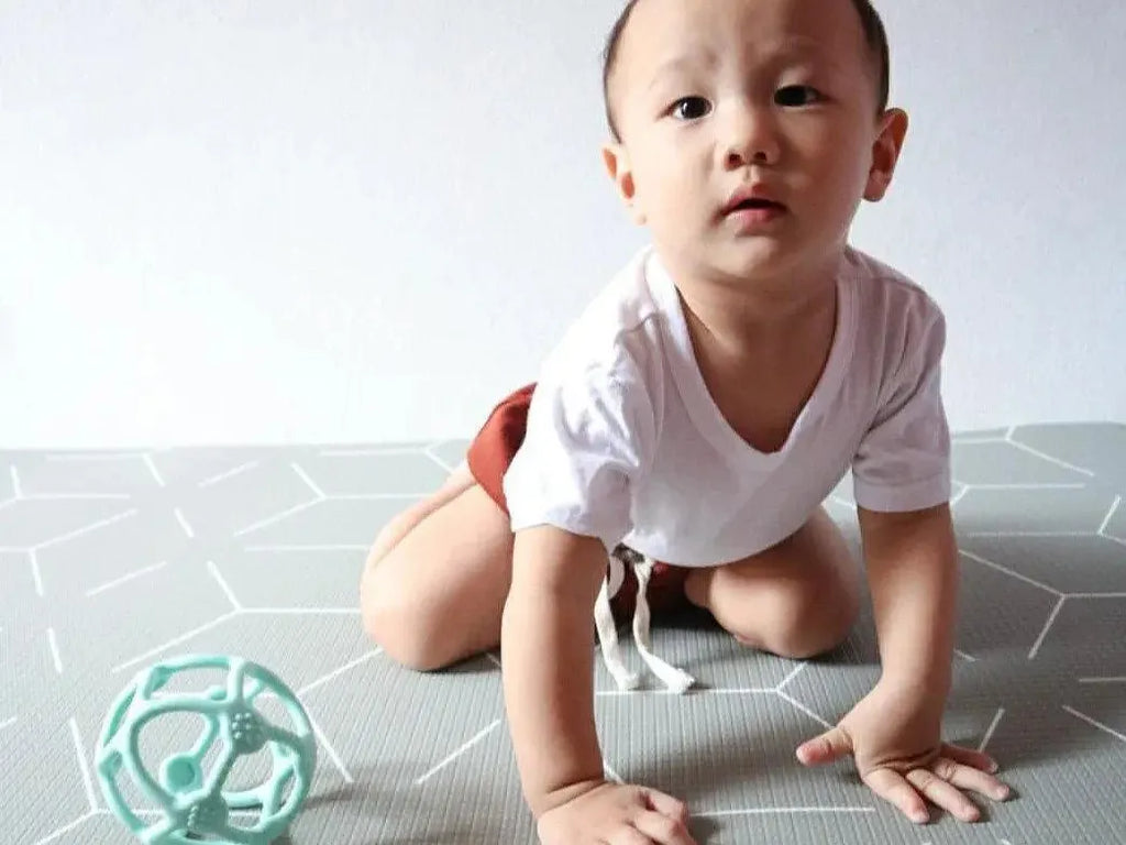 Baby crawling on a patterned floor mat near a teal-colored toy.