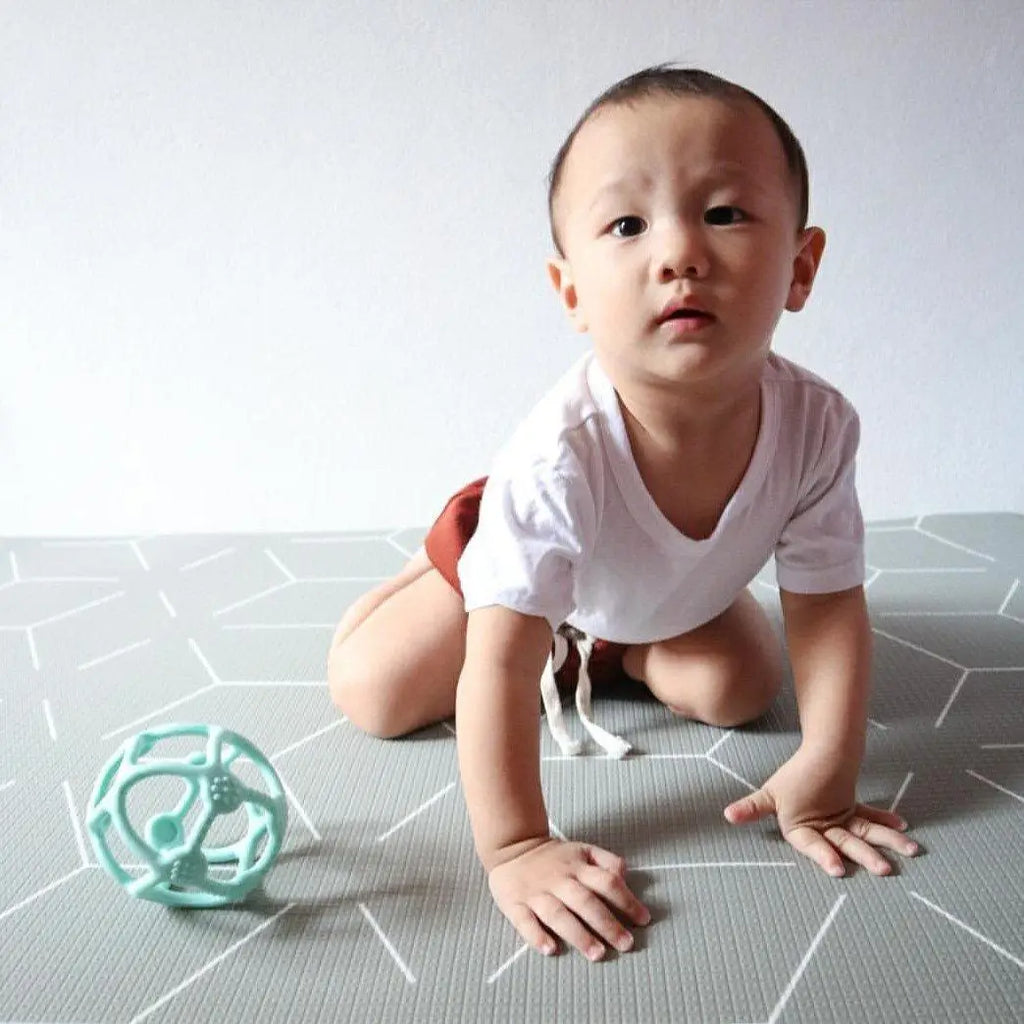 Baby crawling on a patterned floor mat near a teal-colored toy.