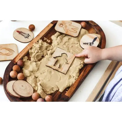 Wooden tray filled with sand or dough, surrounded by various wooden shapes and tools for play or learning.