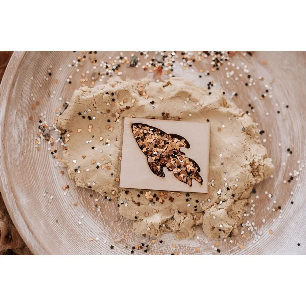 Wooden block with a leaf-shaped cutout filled with mixed seeds and spices atop a bowl of dough.