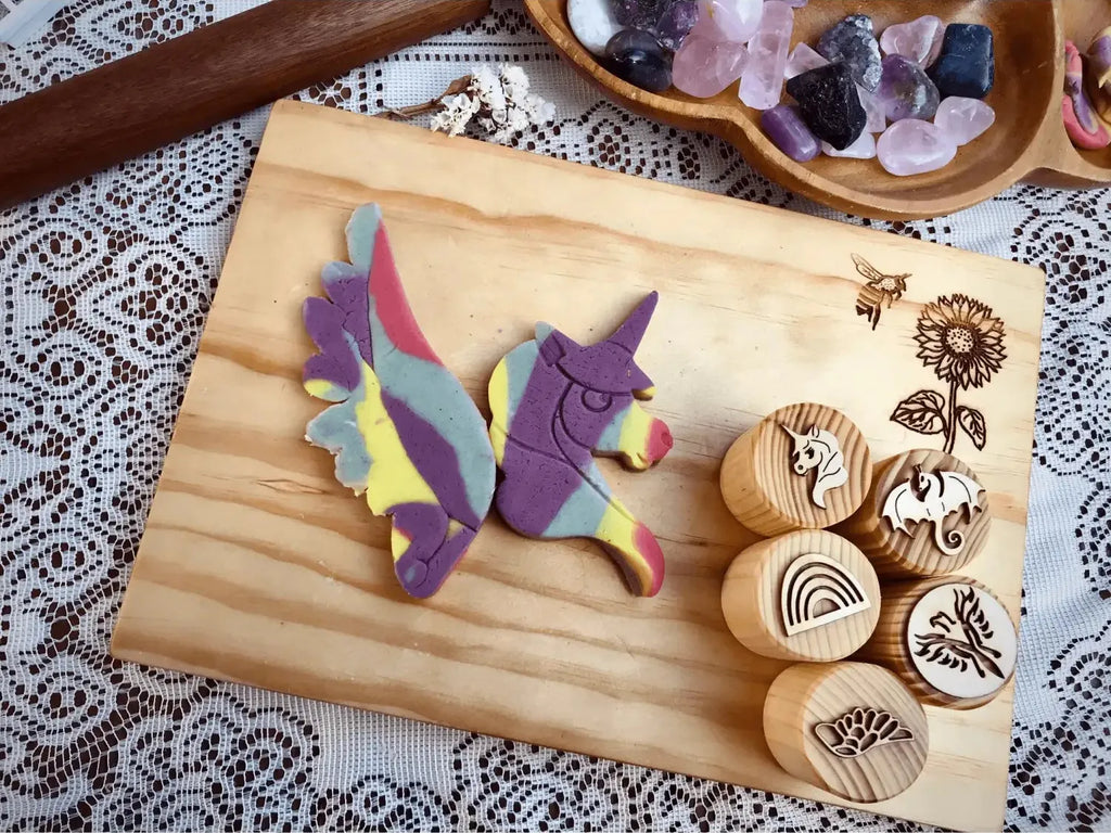 Wooden cutting board displaying colorful clay unicorn figures and round wooden stamps with nature designs.