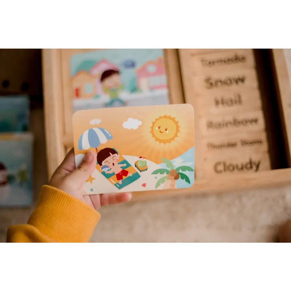 Colorful wooden card depicting a beach scene with a smiling sun and a child relaxing under an umbrella.